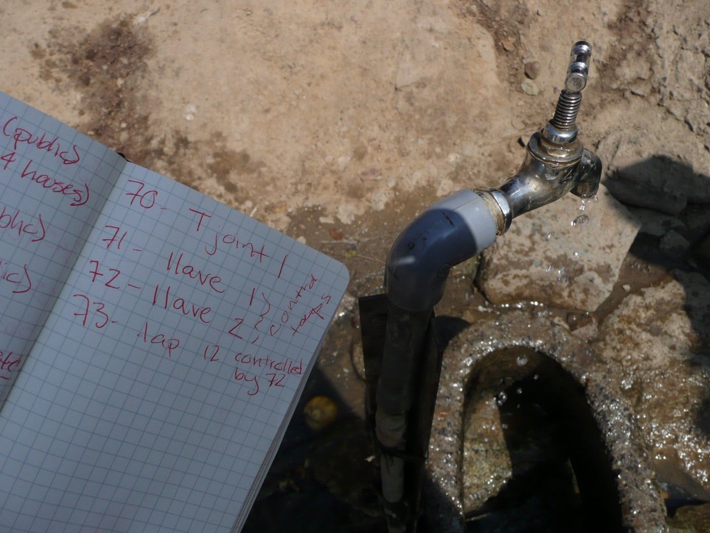 Open field notebook with handwritten numbered survey entries, beside a running metal tap on a plastic riser set into stony ground